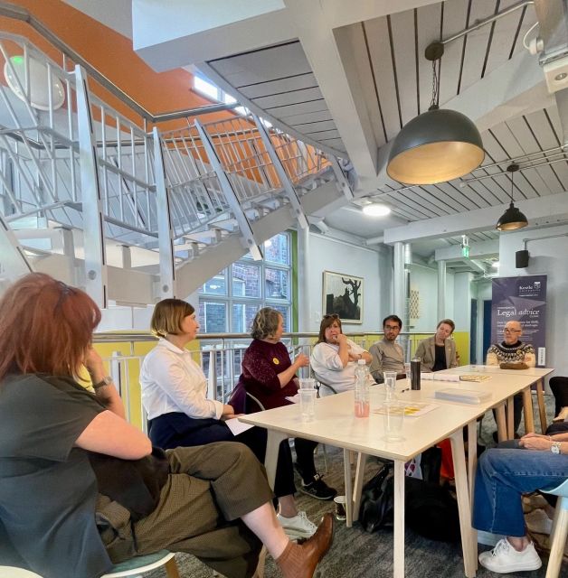 A roundtable discussion featuring Lisa Blower, Helen Burton, Ceri Morgan, Rose Hunter, several guests, and Mike Jackson, co-founder of Lesbians and Gays Support the Miners (LGSM), all seated around a table engaged in conversation.